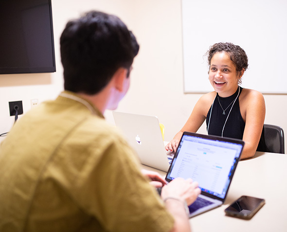 Two students talk over their laptops in a study room