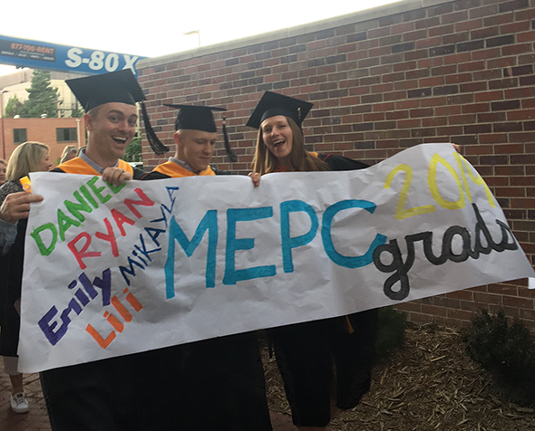 Three grad students in cap and gown walk to their 2019 commencement ceremony across campus holding an MEPC Grads banner