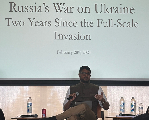 Dr. Debak Das sits with a microphone in hand underneath a projection screen reading "Russia's War on Ukraine: Two Years Since the Full-Scale Invasion"