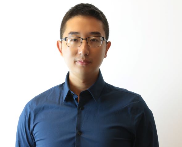 A man wearing a blue shirt and glasses stands in front of a white background