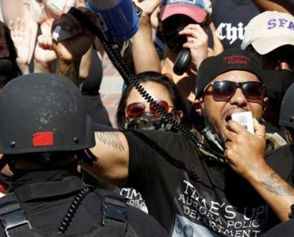 A group of people stand in protest against a line of police in riot gear. One man near the front is speaking into a bullhorn.
