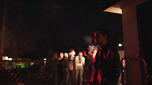 Party Quest film poster. A man stands in the foreground outside at night, looking into the distance. A group of people stand facing away from him in the background, lit by a porch light. . 
