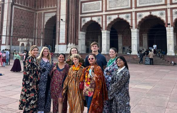 A group of students, teachers, and guides stand in a stone pavilion in front of a building with many arches and columns.