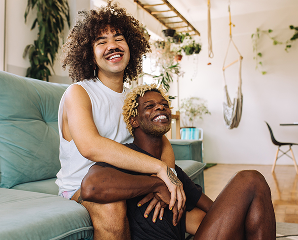 One man sits on a couch, embracing a second man who is sitting in front of him on the floor. The two are smiling comfortably and affectionately.